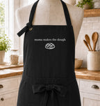 A black kitchen apron displayed on a beige mannequin torso in a bright kitchen setting. The apron has white text reading 'mama makes the dough' above a small white bread illustration, and a waist tie with a bow.