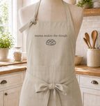 A beige kitchen apron displayed on a mannequin torso in a bright, real kitchen. The apron has the phrase 'mama makes the dough' and a simple bread illustration printed on the chest.