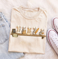 A folded cream-colored t-shirt with a cute graphic of five mice dressed as chefs on a wooden shelf, styled on a white faux fur background with light-wash jeans and white sneakers.