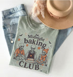 A folded light sage green graphic T-shirt with the words 'Midnight baking CLUB' and a Halloween-themed illustration of three skeletons baking, styled on a white surface with light-wash jeans, a tan hat, and dried grass.