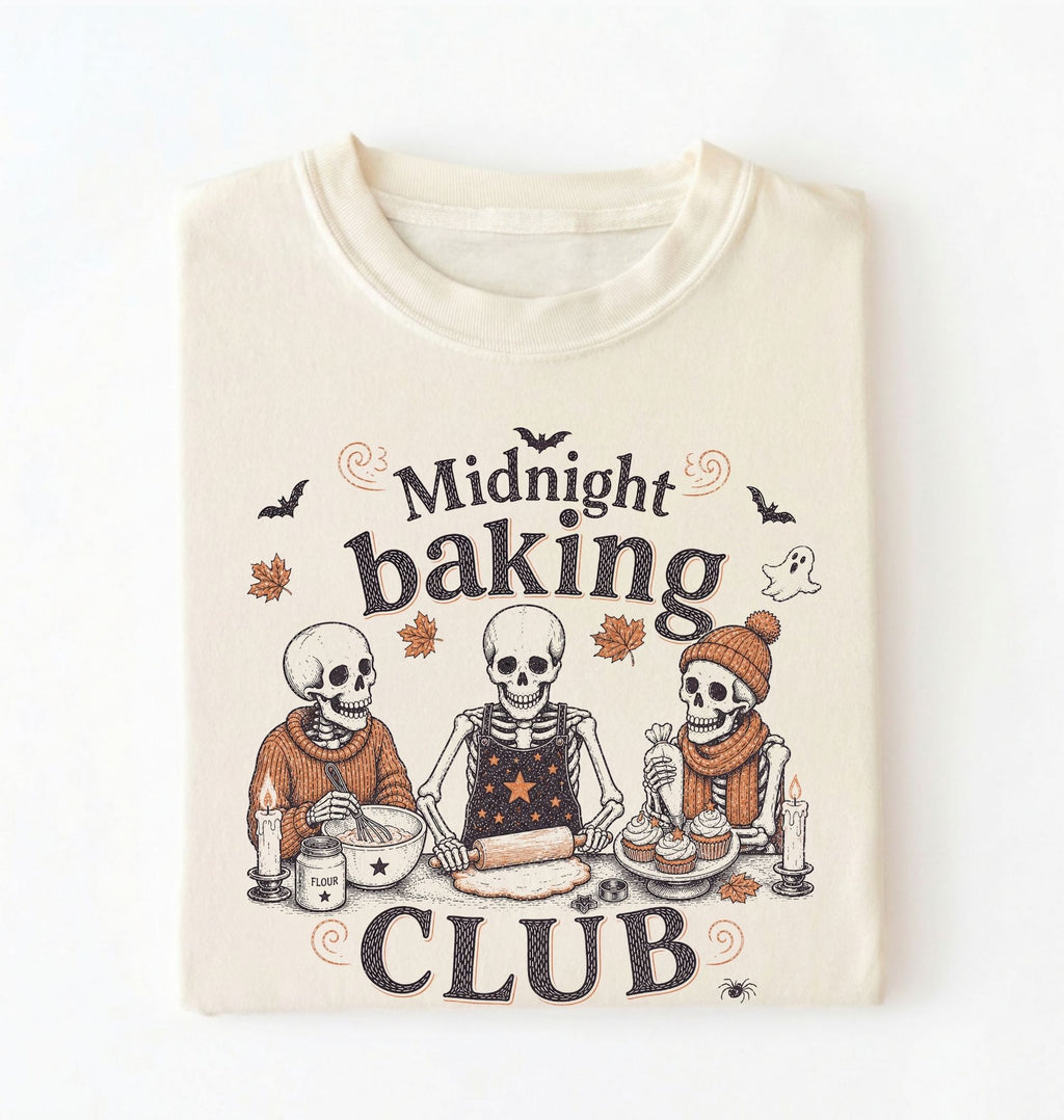 A folded cream-colored t-shirt with a graphic print of three baking skeletons and the text 'Midnight baking CLUB'.