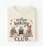 A folded cream-colored t-shirt with a graphic print of three baking skeletons and the text 'Midnight baking CLUB'.