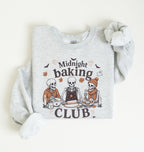 A light heather gray long-sleeve sweatshirt with a cozy rolled cuff, laid flat on a warm off-white background. The front features a graphic of three cartoon skeletons baking together with the text 'Midnight baking CLUB' and small autumn-themed illustrations.