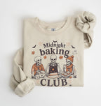 A beige crewneck sweatshirt featuring a 'Midnight Baking Club' Halloween graphic with three baking skeletons, styled flat on a white background with a matching beige knit scrunchie.