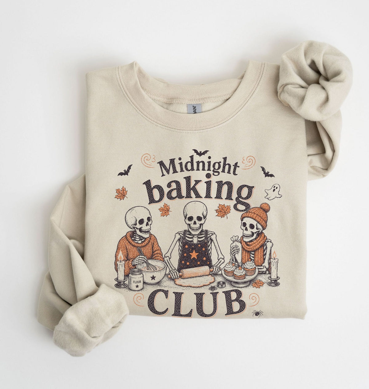 A beige crewneck sweatshirt featuring a 'Midnight Baking Club' Halloween graphic with three baking skeletons, styled flat on a white background with a matching beige knit scrunchie.