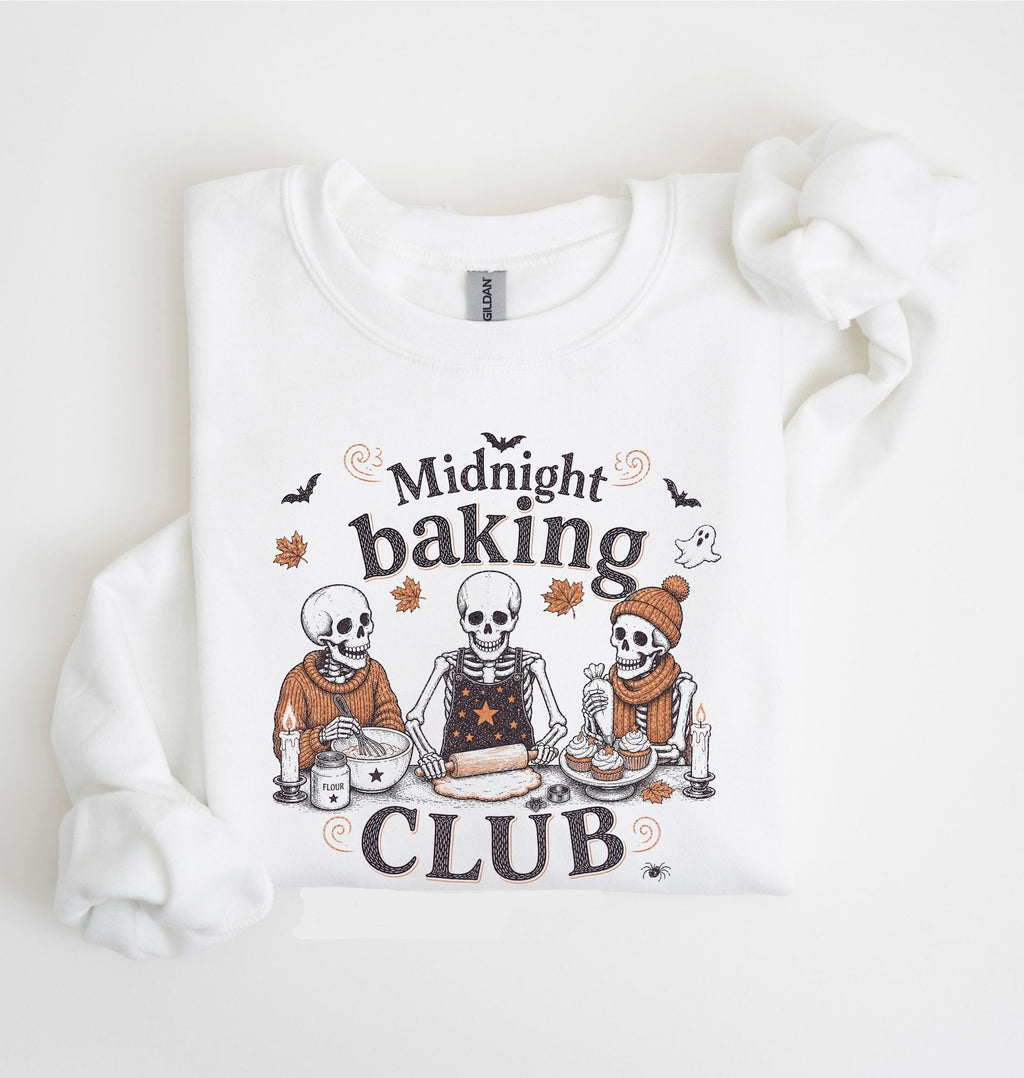 A white long-sleeve sweatshirt with a graphic print of three cartoon skeletons baking together under the words 'Midnight baking CLUB'. The sweatshirt is laid flat on a white background with a matching white fabric scrunchie on the right side.