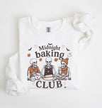A white long-sleeve sweatshirt with a graphic print of three cartoon skeletons baking together under the words 'Midnight baking CLUB'. The sweatshirt is laid flat on a white background with a matching white fabric scrunchie on the right side.