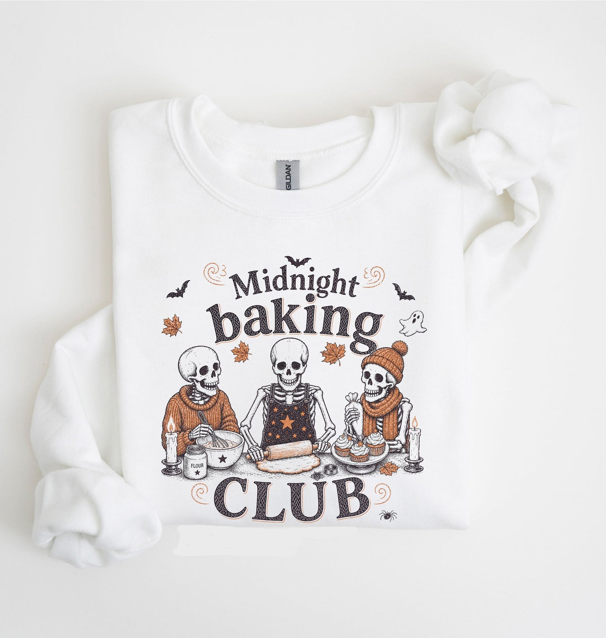A white long-sleeve sweatshirt with a graphic print of three cartoon skeletons baking together under the words 'Midnight baking CLUB'. The sweatshirt is laid flat on a white background with a matching white fabric scrunchie on the right side.