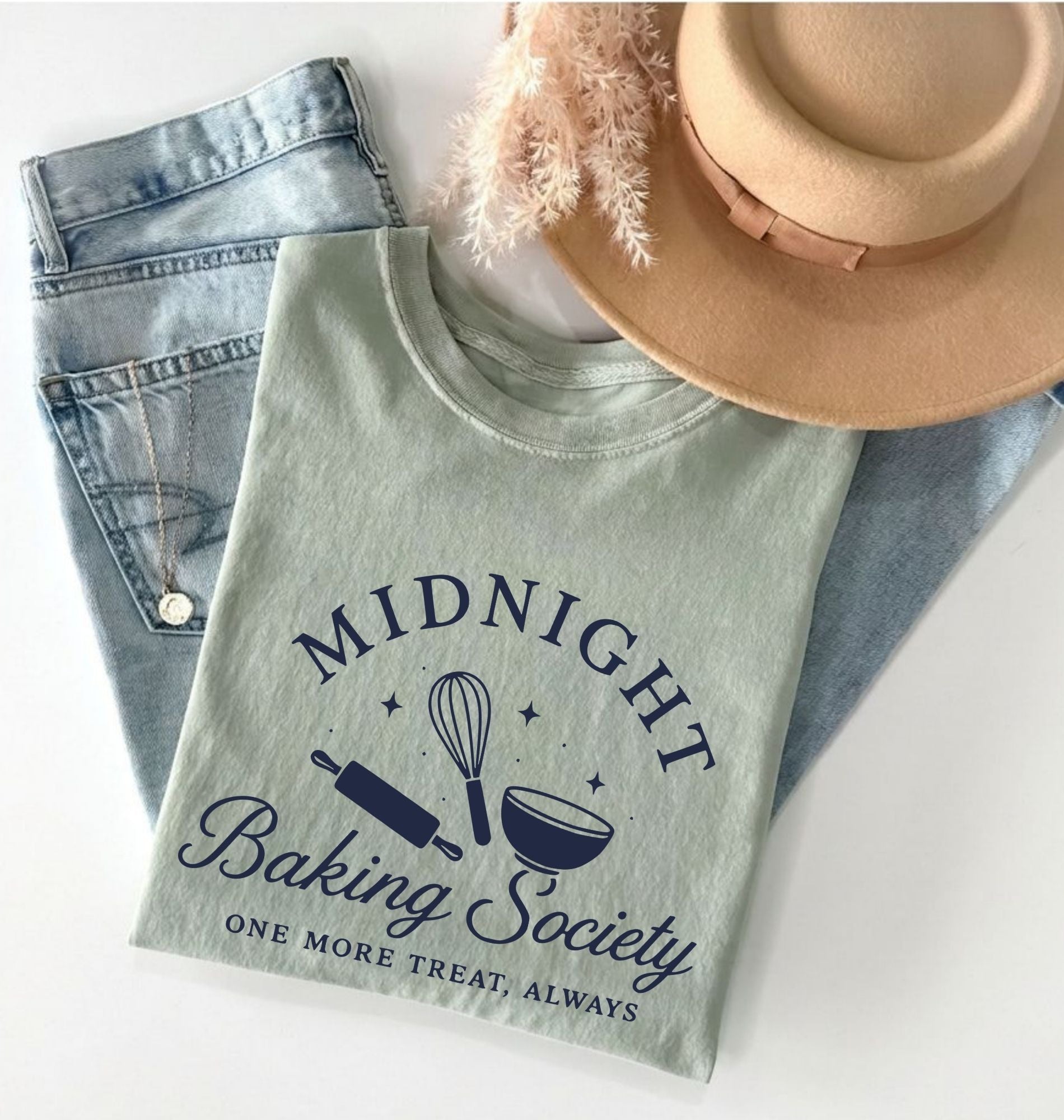 A folded sage green graphic T-shirt with the text 'MIDNIGHT Baking Society' and baking-themed illustrations, styled on top of light blue jeans and a beige hat on a white background.