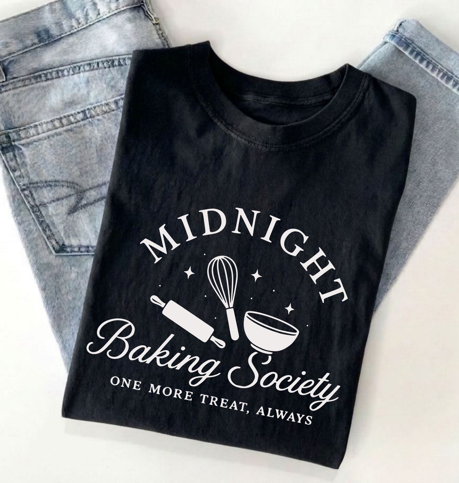 A black short-sleeve T-shirt laid flat on a white surface with light blue jeans partially visible behind it. The shirt has a white graphic print reading “MIDNIGHT Baking Society” with baking tools and the phrase “ONE MORE TREAT, ALWAYS.