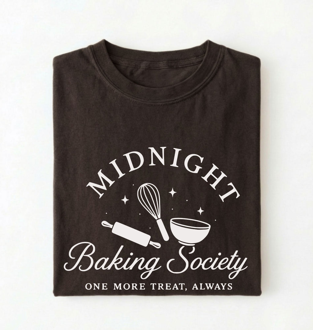 A folded dark brown t-shirt with a white graphic print that reads 'MIDNIGHT Baking Society' and 'ONE MORE TREAT, ALWAYS' with baking utensil illustrations, shown on a plain white background.