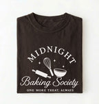 A folded dark brown t-shirt with a white graphic print that reads 'MIDNIGHT Baking Society' and 'ONE MORE TREAT, ALWAYS' with baking utensil illustrations, shown on a plain white background.