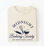 A folded cream-colored t-shirt with a navy blue graphic that reads 'MIDNIGHT Baking Society' and 'ONE MORE TREAT, ALWAYS' with baking utensil illustrations, shown on a plain white background.