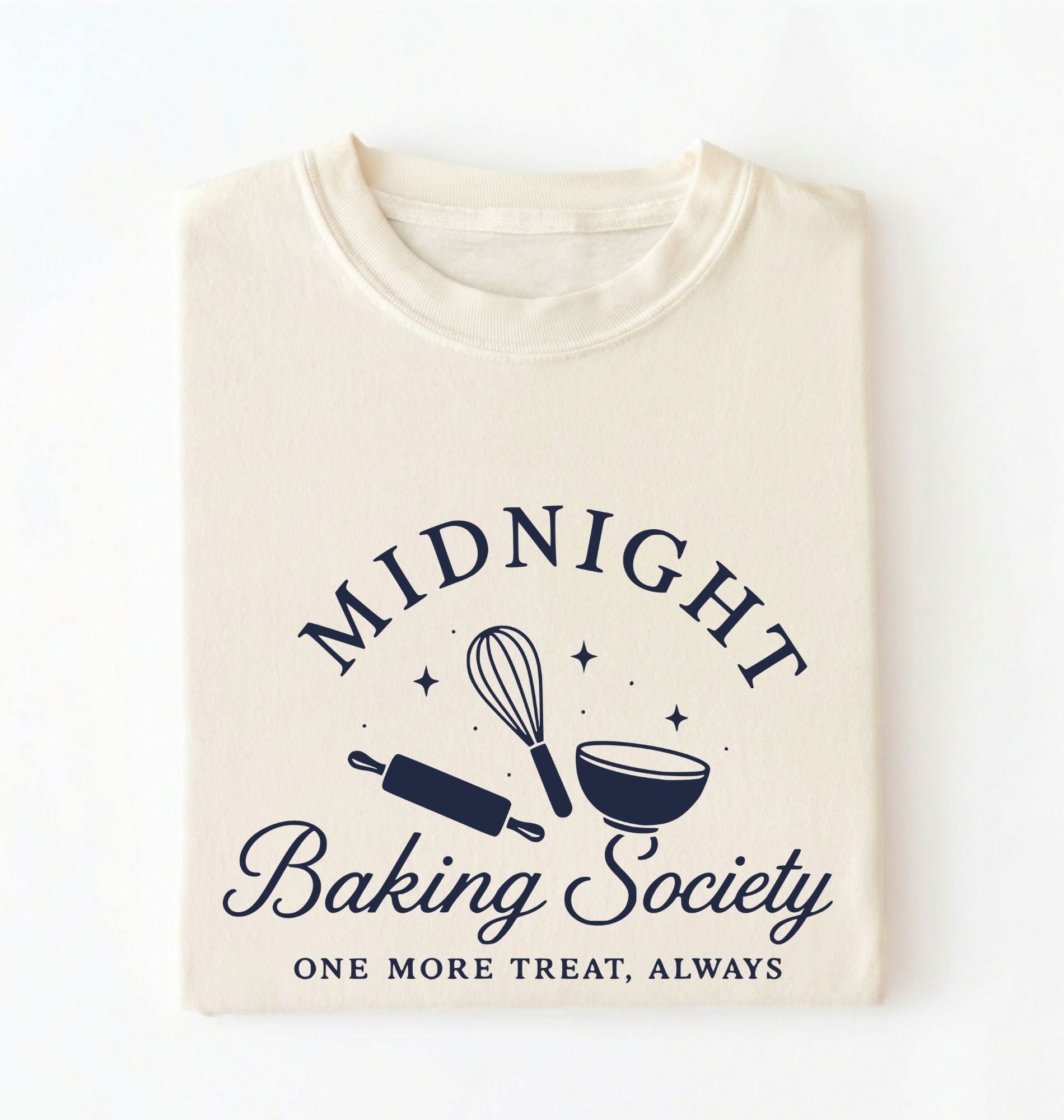 A folded cream-colored t-shirt with a navy blue graphic that reads 'MIDNIGHT Baking Society' and 'ONE MORE TREAT, ALWAYS' with baking utensil illustrations, shown on a plain white background.