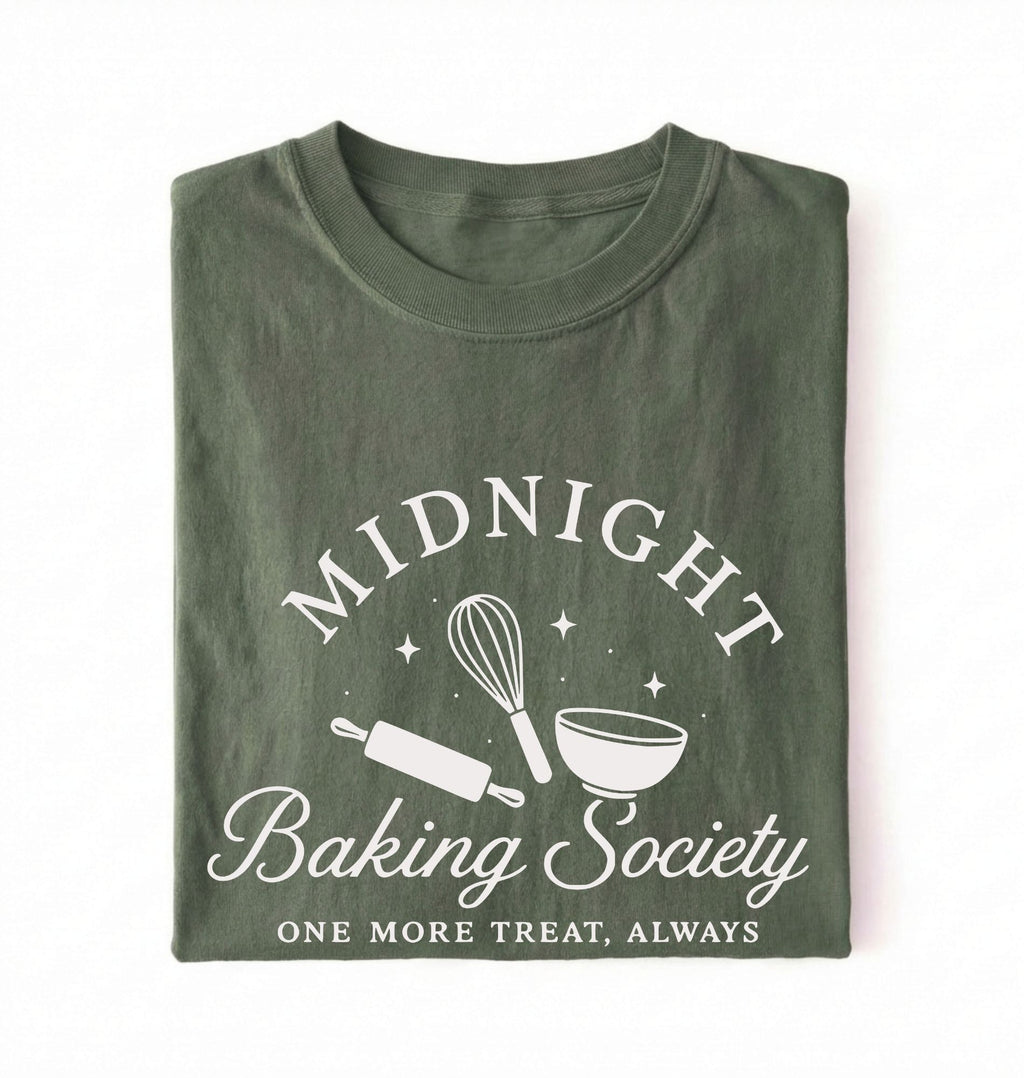 A folded dark olive green t-shirt with a white graphic print that reads 'MIDNIGHT Baking Society' and 'ONE MORE TREAT, ALWAYS' with illustrations of a whisk, rolling pin, and bowl on a white background
