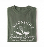 A folded dark olive green t-shirt with a white graphic print that reads 'MIDNIGHT Baking Society' and 'ONE MORE TREAT, ALWAYS' with illustrations of a whisk, rolling pin, and bowl on a white background