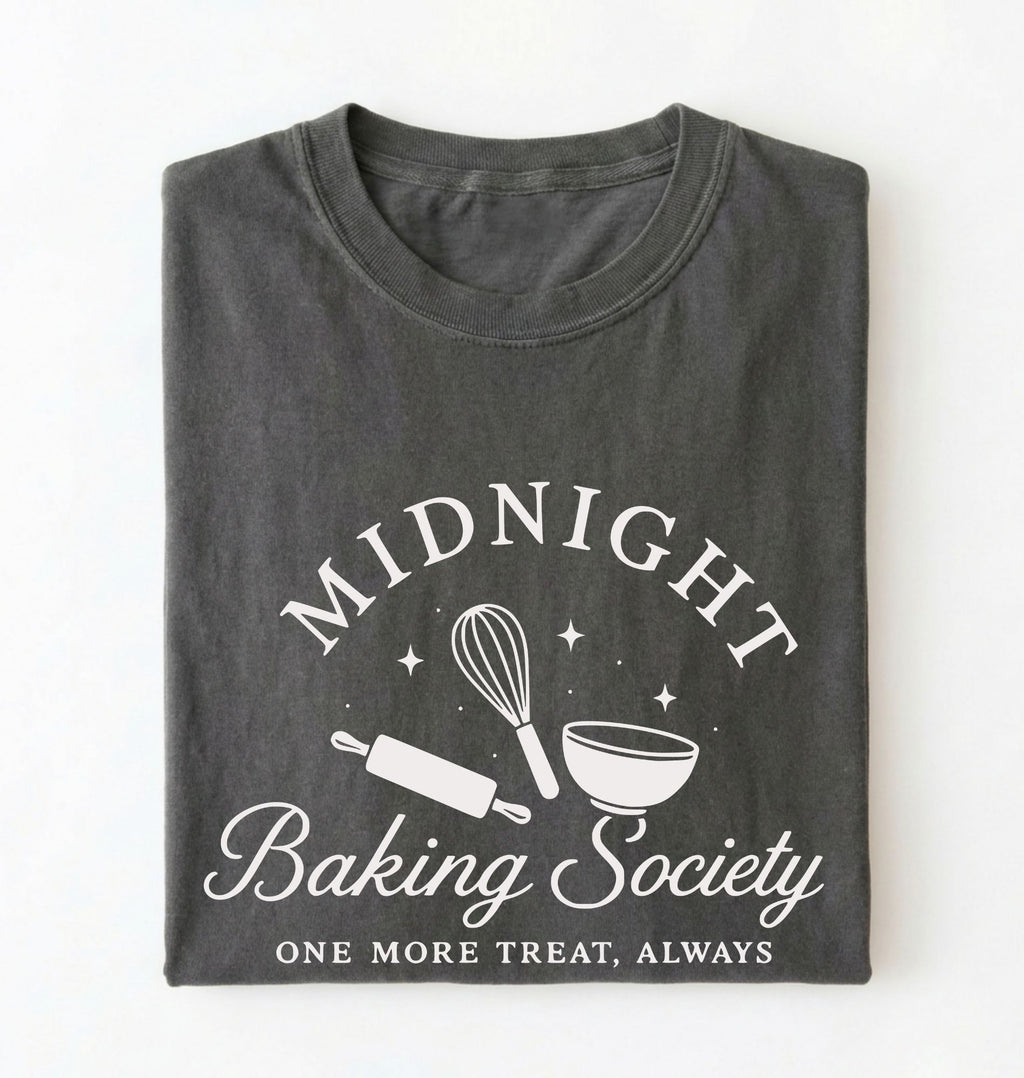 A dark gray folded t-shirt with a white graphic print that reads 'MIDNIGHT Baking Society' and 'ONE MORE TREAT, ALWAYS' with baking tools like a whisk, rolling pin, and bowl.