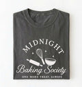A dark gray folded t-shirt with a white graphic print that reads 'MIDNIGHT Baking Society' and 'ONE MORE TREAT, ALWAYS' with baking tools like a whisk, rolling pin, and bowl.