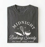 A dark gray folded t-shirt with a white graphic print that reads 'MIDNIGHT Baking Society' and 'ONE MORE TREAT, ALWAYS' with baking tools like a whisk, rolling pin, and bowl.