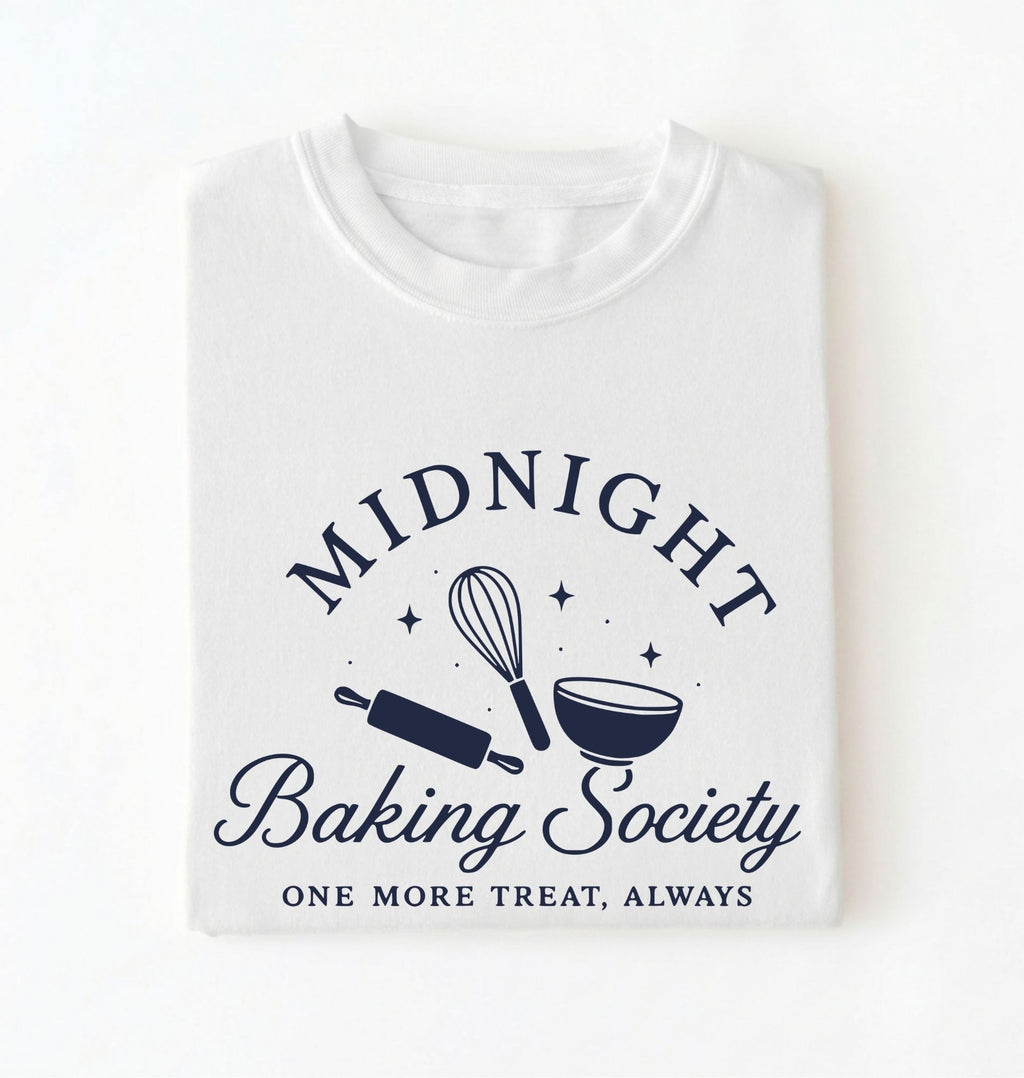 A folded white t-shirt with a navy blue graphic that reads 'MIDNIGHT Baking Society' and 'ONE MORE TREAT, ALWAYS' with illustrations of a whisk, rolling pin, and bowl.