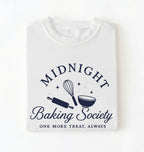 A folded white t-shirt with a navy blue graphic that reads 'MIDNIGHT Baking Society' and 'ONE MORE TREAT, ALWAYS' with illustrations of a whisk, rolling pin, and bowl.