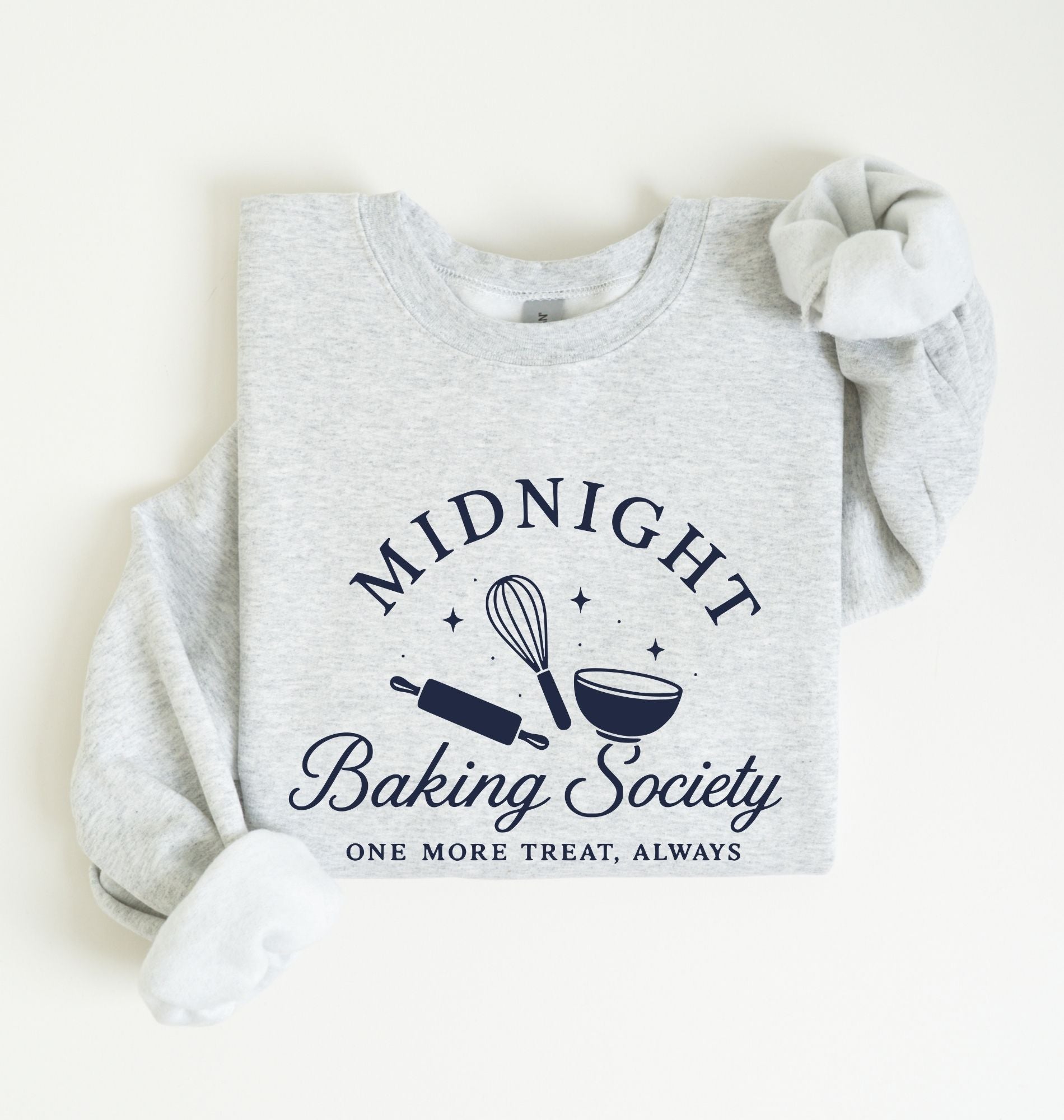 A light heather gray long-sleeve sweatshirt laid flat on a cream background, featuring a navy blue graphic that reads 'MIDNIGHT Baking Society' with baking tools and the phrase 'ONE MORE TREAT, ALWAYS.'.