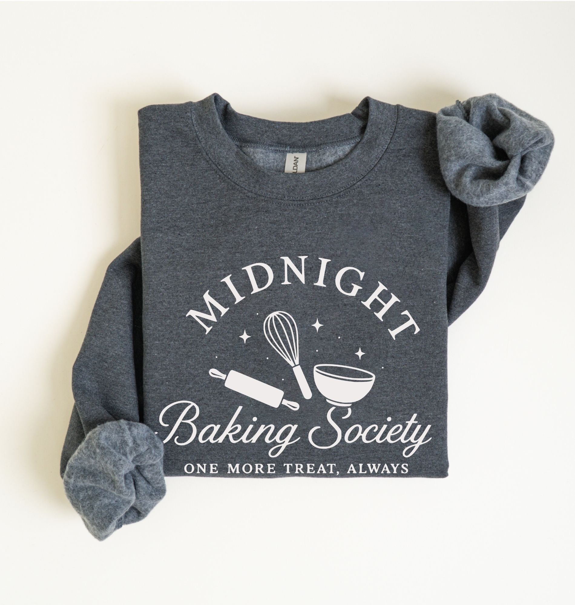 A folded dark heather gray sweatshirt with rolled cuffs, featuring a white graphic print that reads 'MIDNIGHT Baking Society' with a whisk, rolling pin, and bowl illustration, and the phrase 'ONE MORE TREAT, ALWAYS' below.