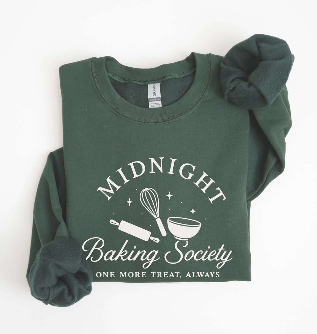A dark green crewneck sweatshirt with rolled sleeves, featuring a white graphic print that reads 'MIDNIGHT Baking Society' and 'ONE MORE TREAT, ALWAYS' with baking tools like a whisk, rolling pin, and bowl. The sweatshirt is displayed flat on a white background with a matching scrunchie.