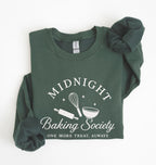 A dark green crewneck sweatshirt with rolled sleeves, featuring a white graphic print that reads 'MIDNIGHT Baking Society' and 'ONE MORE TREAT, ALWAYS' with baking tools like a whisk, rolling pin, and bowl. The sweatshirt is displayed flat on a white background with a matching scrunchie.