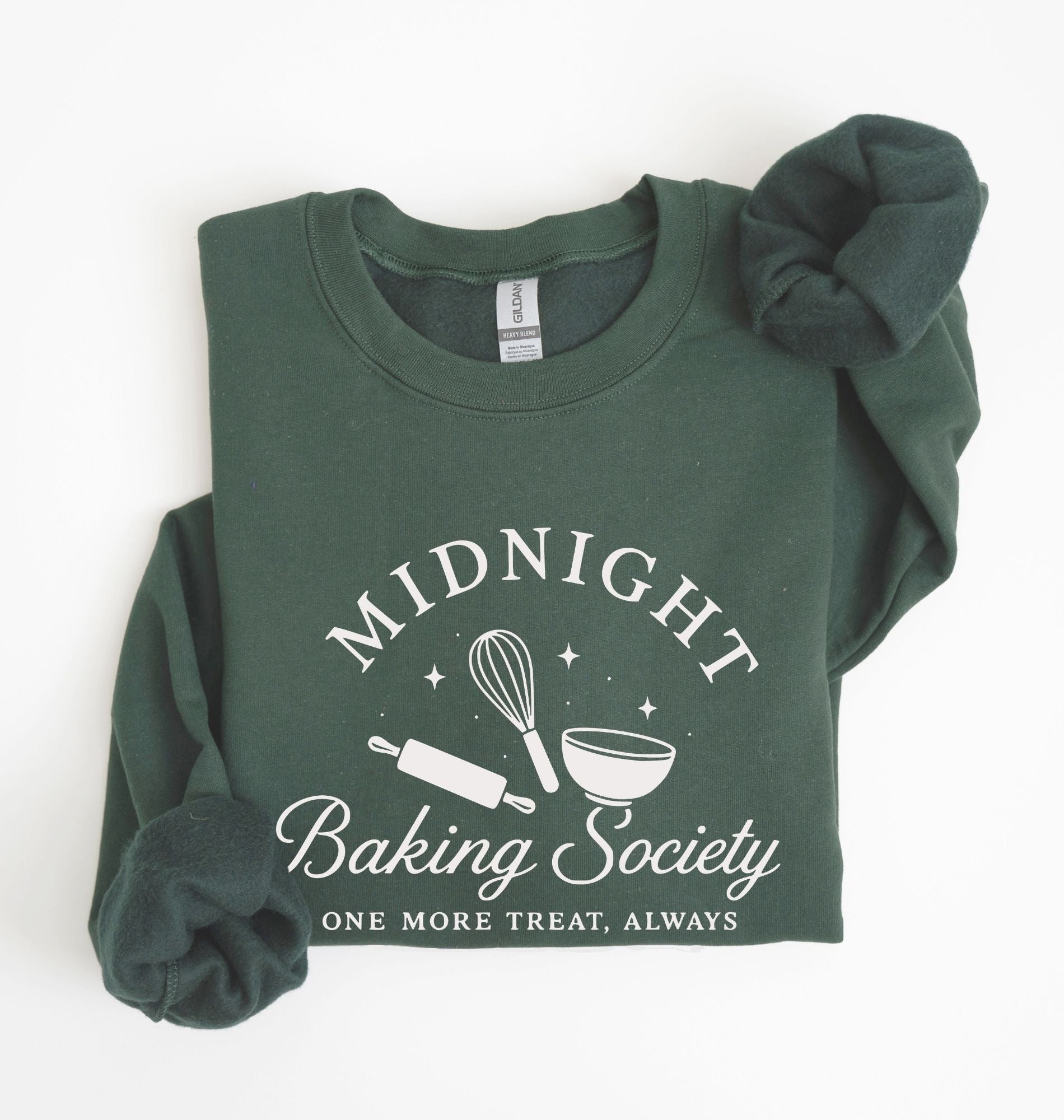 A dark green crewneck sweatshirt with rolled sleeves, featuring a white graphic print that reads 'MIDNIGHT Baking Society' and 'ONE MORE TREAT, ALWAYS' with baking tools like a whisk, rolling pin, and bowl. The sweatshirt is displayed flat on a white background with a matching scrunchie.