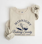 A folded beige crewneck sweatshirt with rolled sleeves, featuring a navy blue graphic that reads 'MIDNIGHT Baking Society' with baking tools and the phrase 'ONE MORE TREAT, ALWAYS'.