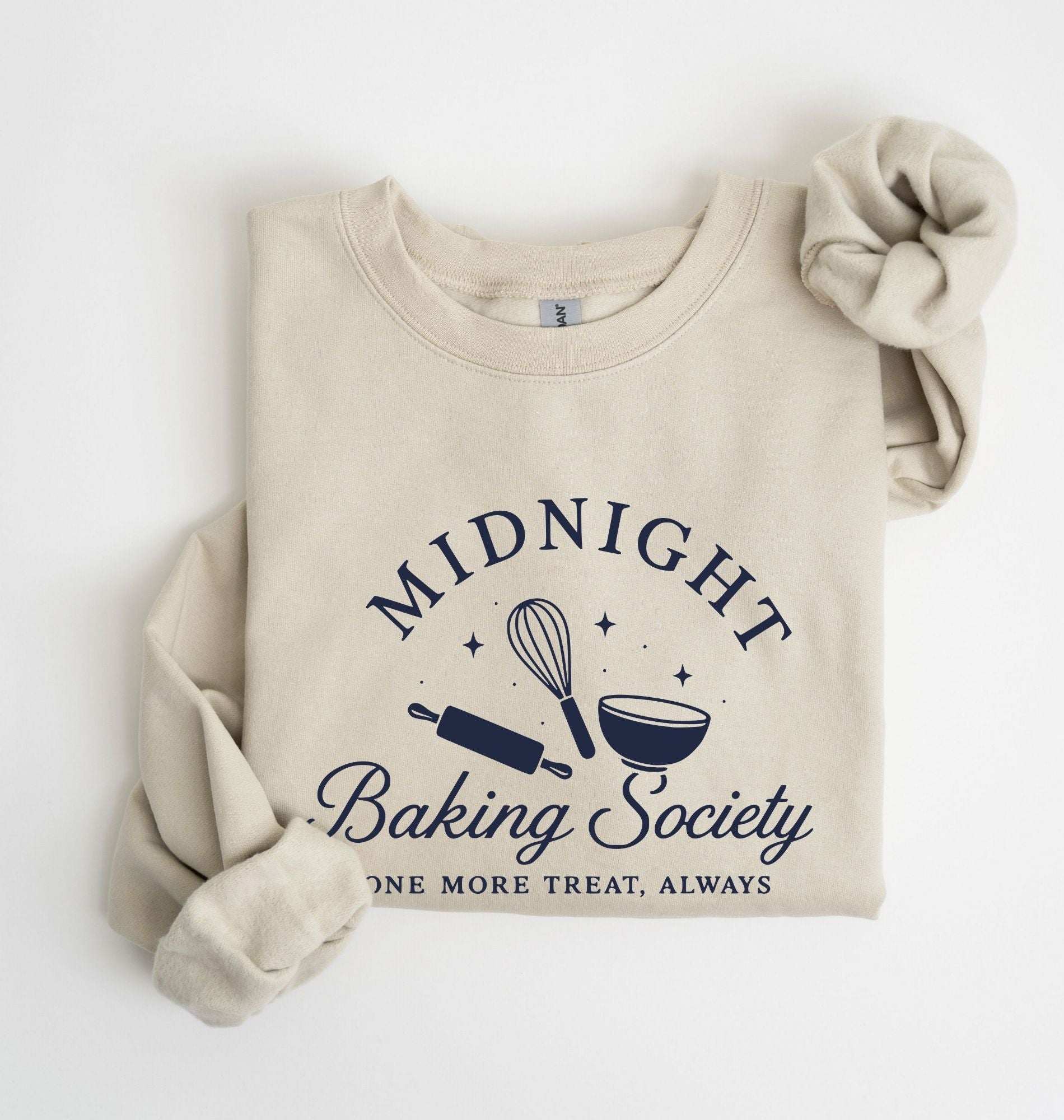 A folded beige crewneck sweatshirt with rolled sleeves, featuring a navy blue graphic that reads 'MIDNIGHT Baking Society' with baking tools and the phrase 'ONE MORE TREAT, ALWAYS'.
