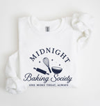 A folded white sweatshirt on a white background, featuring a navy blue graphic that reads 'MIDNIGHT Baking Society' with a whisk, rolling pin, and bowl illustration, and the phrase 'ONE MORE TREAT, ALWAYS' below.