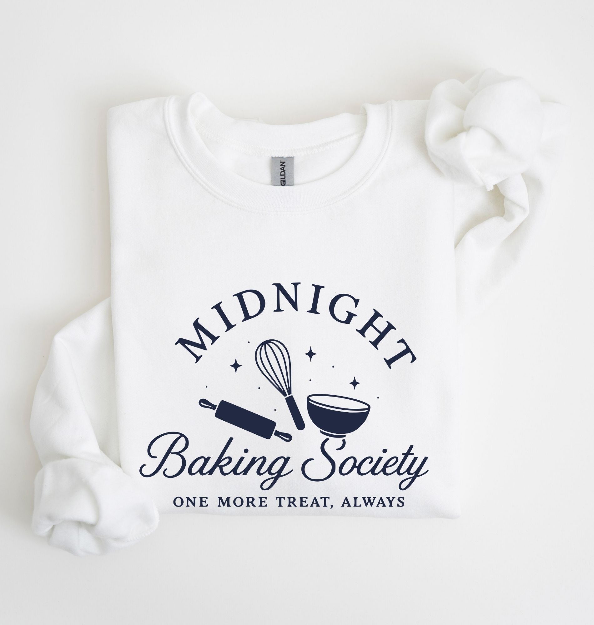 A folded white sweatshirt on a white background, featuring a navy blue graphic that reads 'MIDNIGHT Baking Society' with a whisk, rolling pin, and bowl illustration, and the phrase 'ONE MORE TREAT, ALWAYS' below.