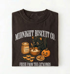 A dark brown folded t-shirt with a Halloween-themed graphic print featuring a black cat, a pumpkin, cookies, and a flour sack with the text 'MIDNIGHT BISCUIT CO. FRESH FROM THE CATACOMBS'.