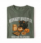 A folded dark olive green t-shirt on a white background, featuring a graphic print of a black cat mixing dough next to a flour sack, pumpkin, and cookies, with the text 'MIDNIGHT BISCUIT CO.' and 'FRESH FROM THE CATACOMBS'.