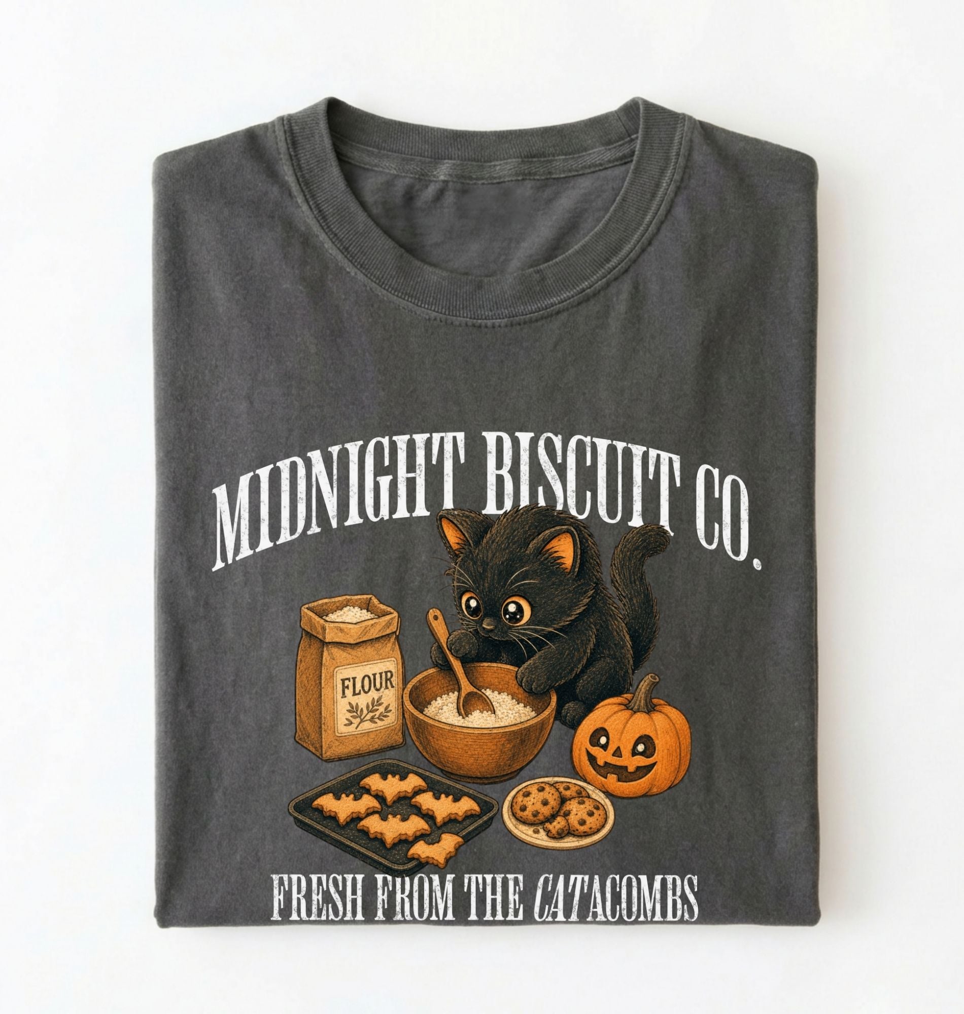 A folded dark gray t-shirt with a graphic print of a black cat baking cookies with flour, a pumpkin, and the text 'MIDNIGHT BISCUIT CO.' and 'FRESH FROM THE CATACOMBS'.