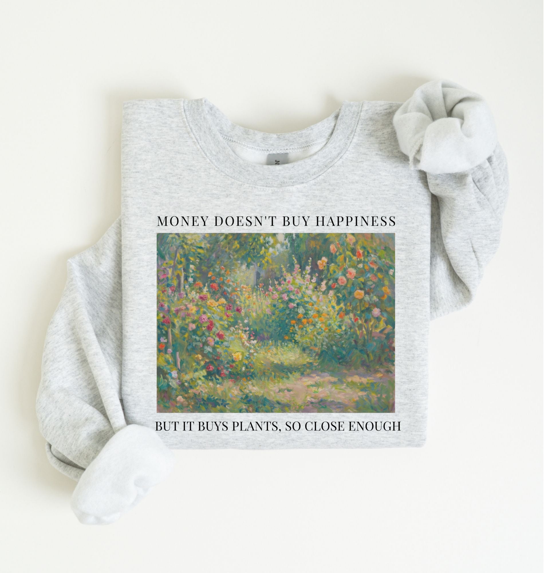 Flat lay of a light gray crewneck sweatshirt on a white background, featuring an impressionist floral garden artwork and black text reading “Money doesn’t buy happiness, but it buys plants, so close enough.