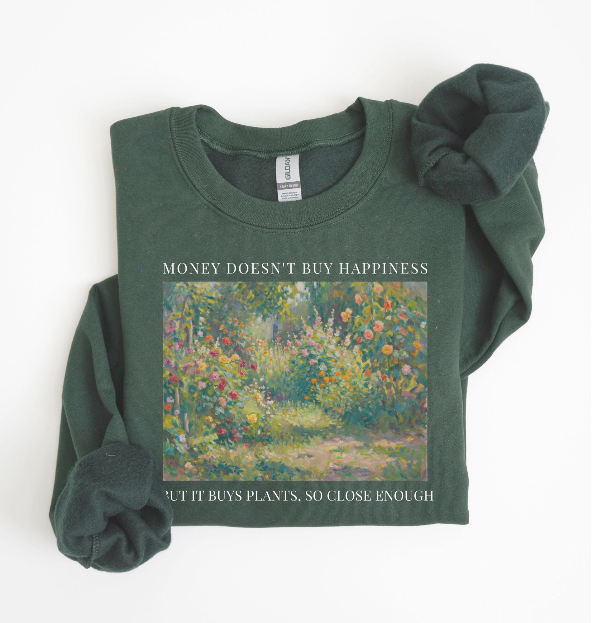 Flat lay of a forest green crewneck sweatshirt on a white background, featuring an impressionist floral garden artwork and black text reading “Money doesn’t buy happiness, but it buys plants, so close enough.”
