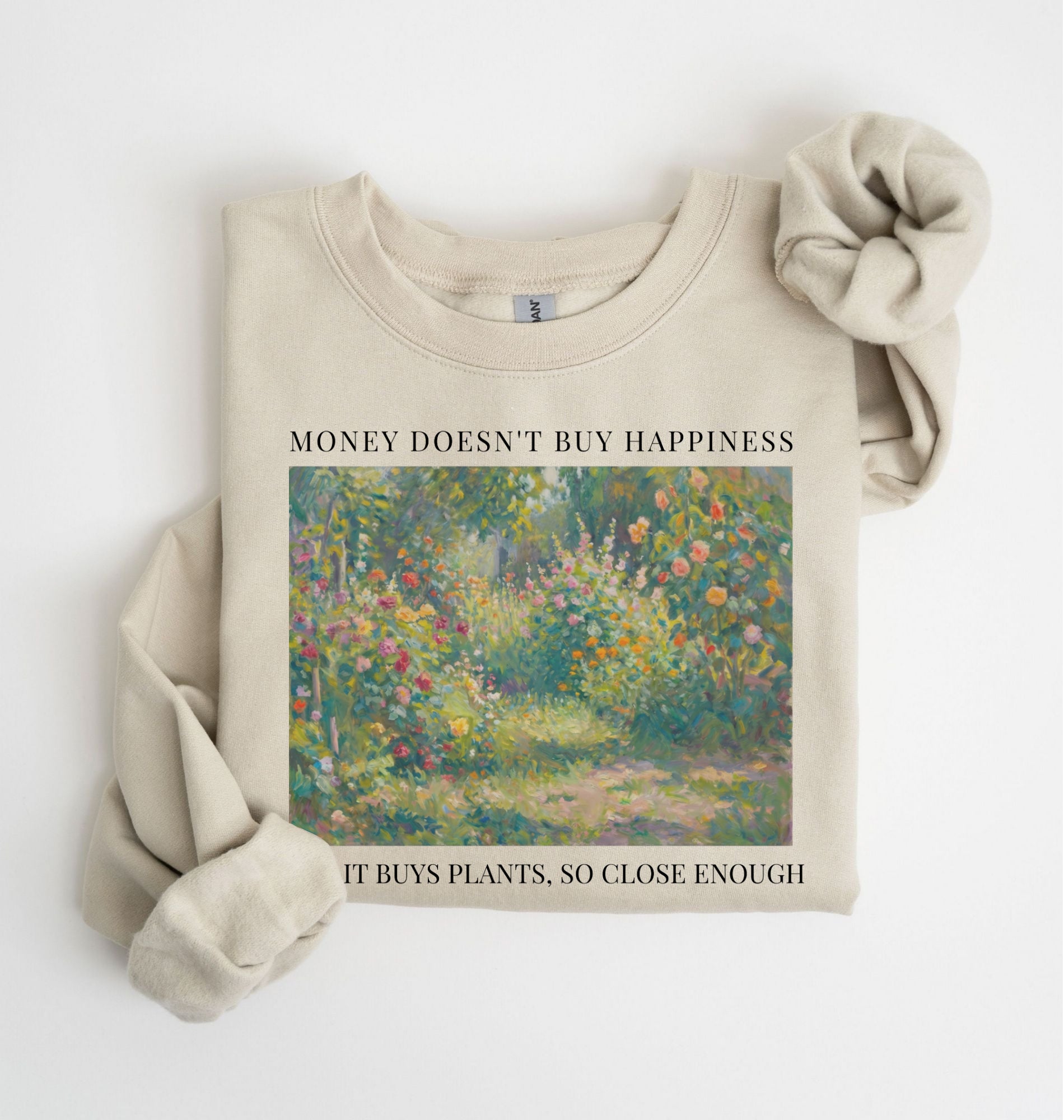 Flat lay of a sand crewneck sweatshirt on a white background, featuring an impressionist floral garden artwork and black text reading “Money doesn’t buy happiness, but it buys plants, so close enough.