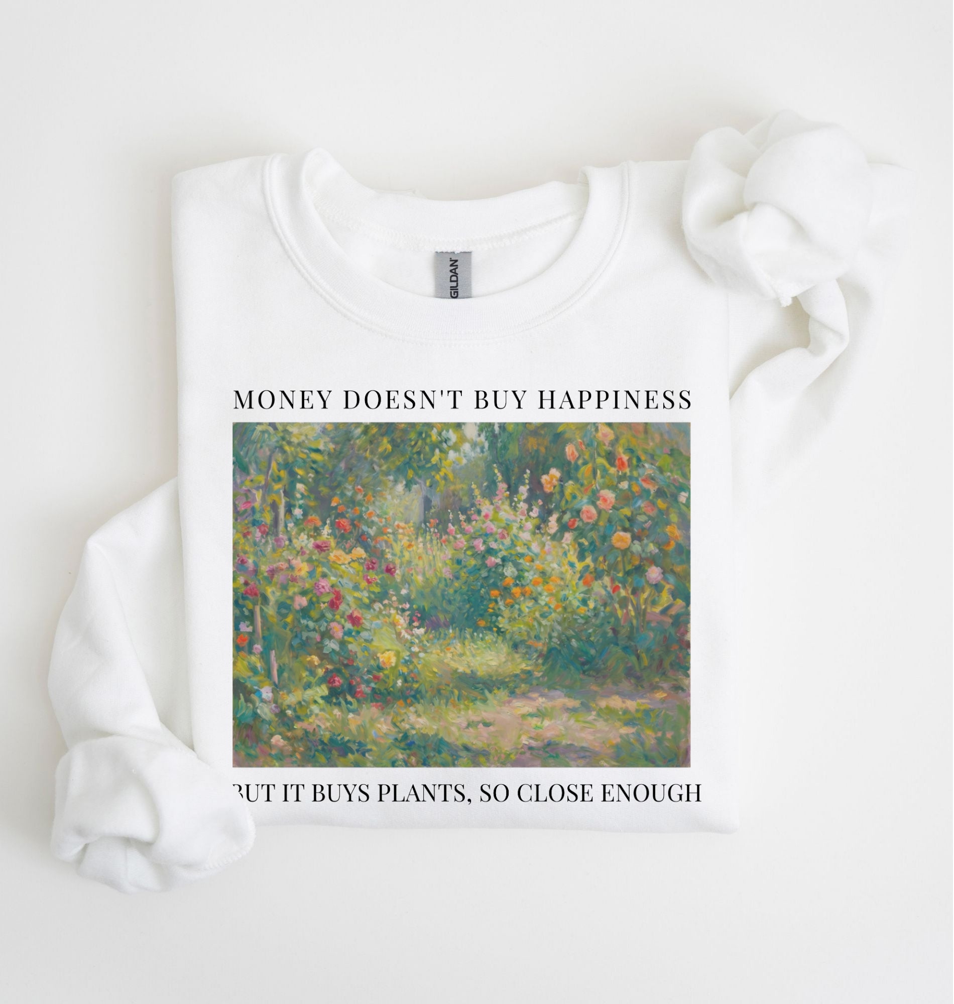 Flat lay of a white crewneck sweatshirt on a white background, featuring an impressionist floral garden artwork and black text reading “Money doesn’t buy happiness, but it buys plants, so close enough.