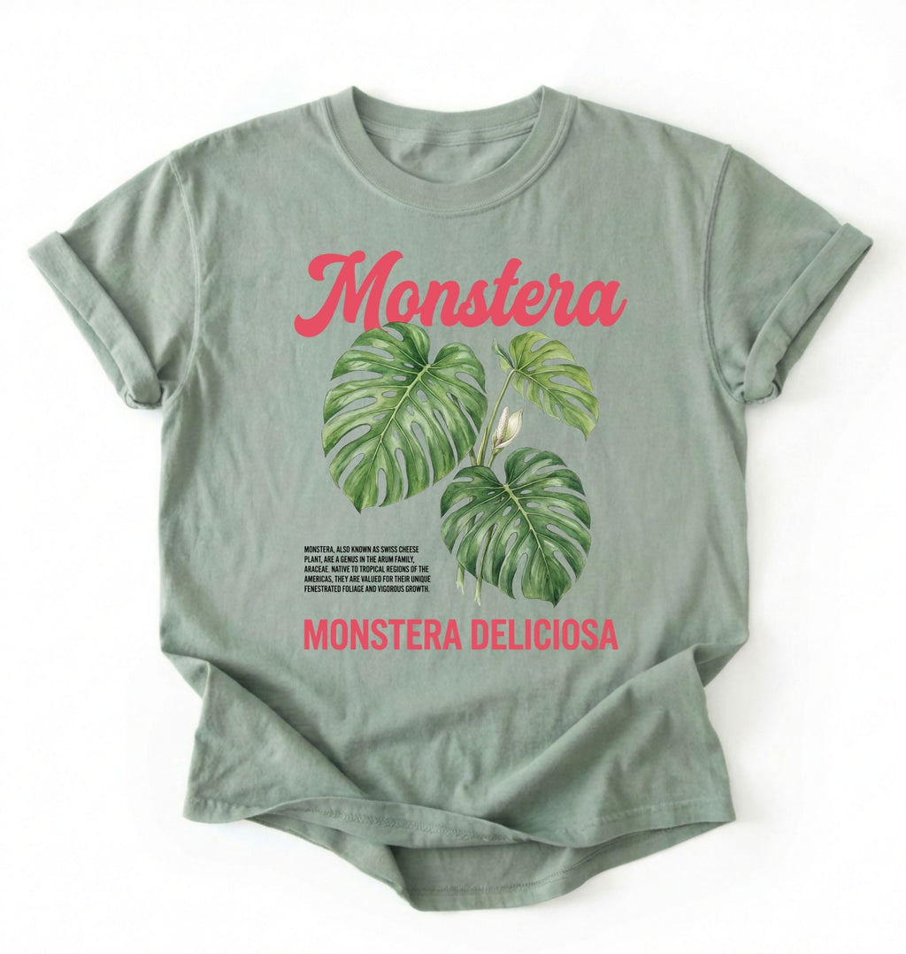 Flat lay of a bay t-shirt featuring a botanical illustration of Monstera leaves with text reading “Monstera Deliciosa.” plant shirt with tropical leaf illustration perfect gift for plant lovers and houseplant enthusiasts on a white background