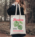 Person holding a tote bag with 'Monstera' design in a forest setting