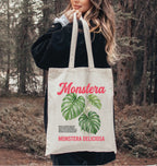 Person holding a tote bag with 'Monstera' design in a forest setting
