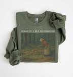Flat lay of an military green Cottagecore mushroom sweatshirt Hold Up I See Mushrooms design beige crewneck for nature lovers on a white background