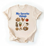 Flat lay of a cream my favorite things shirt with bread chicken coffee plants and cottagecore homestead illustrations on a white background