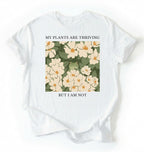 Flat lay of a white Gardener t-shirt with floral design and text my plants are thriving but i am not plant lover gift on a white background 