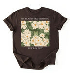 Flat lay of a dark_chocolate Gardener t-shirt with floral design and text my plants are thriving but i am not plant lover gift on a white background 