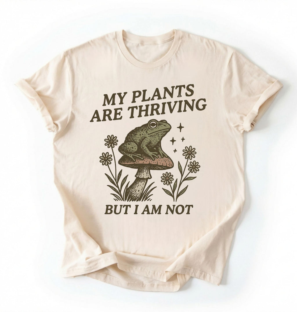 Flat lay of an ivory Comfort Colors sweatshirt with frog on mushroom illustration and text my plants are thriving but I am not on a white background