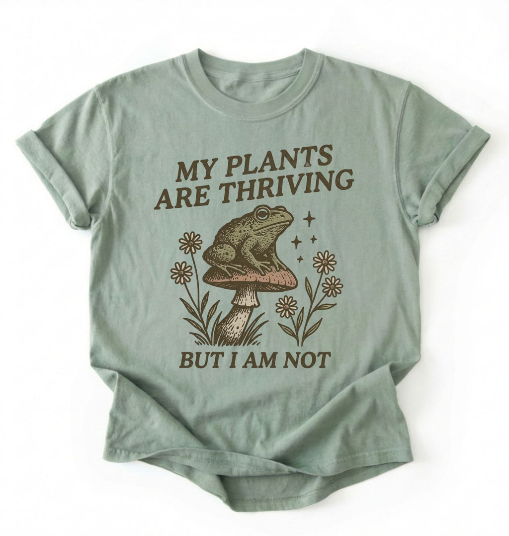 Flat lay of a bay sweatshirt with frog on mushroom illustration and text my plants are thriving but I am not on a white background